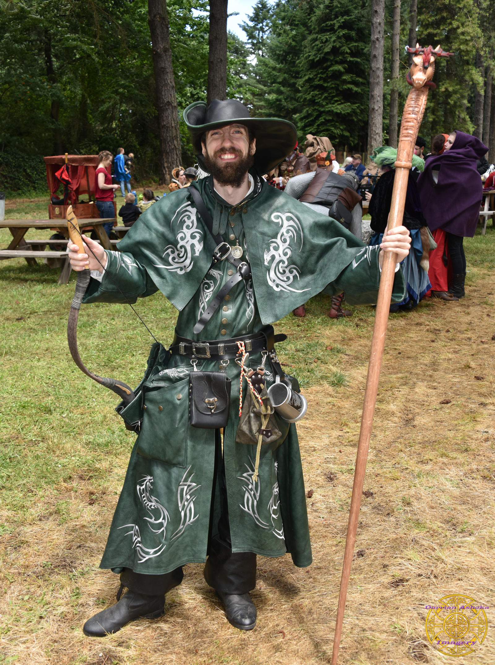 a smiling man in a meticulously crafted green leather medieval costume stands proudly at a renaissance festival. his attire includes a widebrimmed hat, a long coat adorned with silver dragonlike designs, a quiver, bow and arrow, and a staff topped with a carved dragon head.  he is surrounded by the sights and sounds of the festival, with other costumed individuals and picnic tables visible in the background.  the setting appears to be an outdoor woodland area.