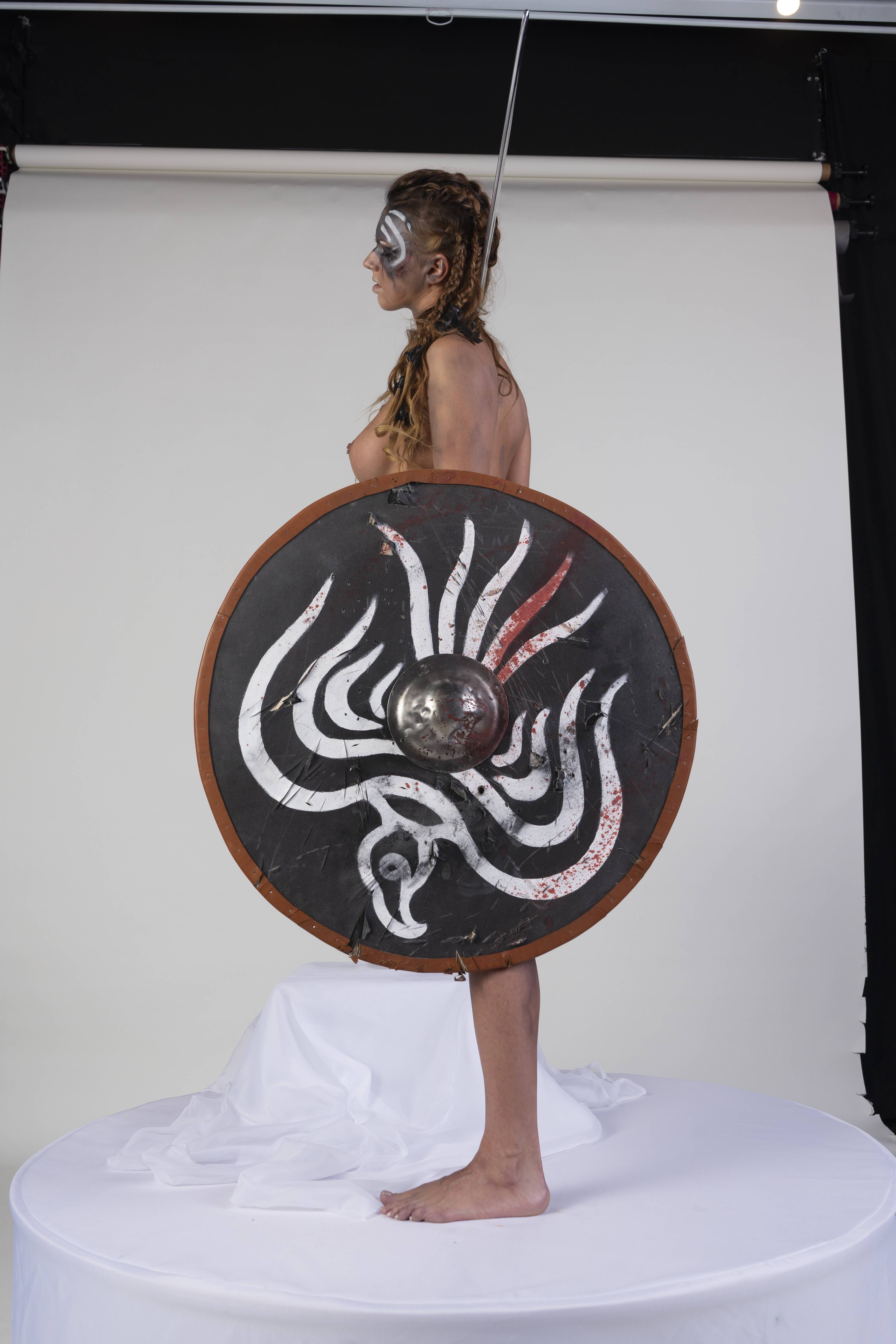 a woman with bare breasts stands facing left with a round shield in front of her torso, holding a sword behind her. she has dark makeup around her eyes and braids in her hair. the shield is black with white and red decorations. she is standing on a white platform in front of a white backdrop.