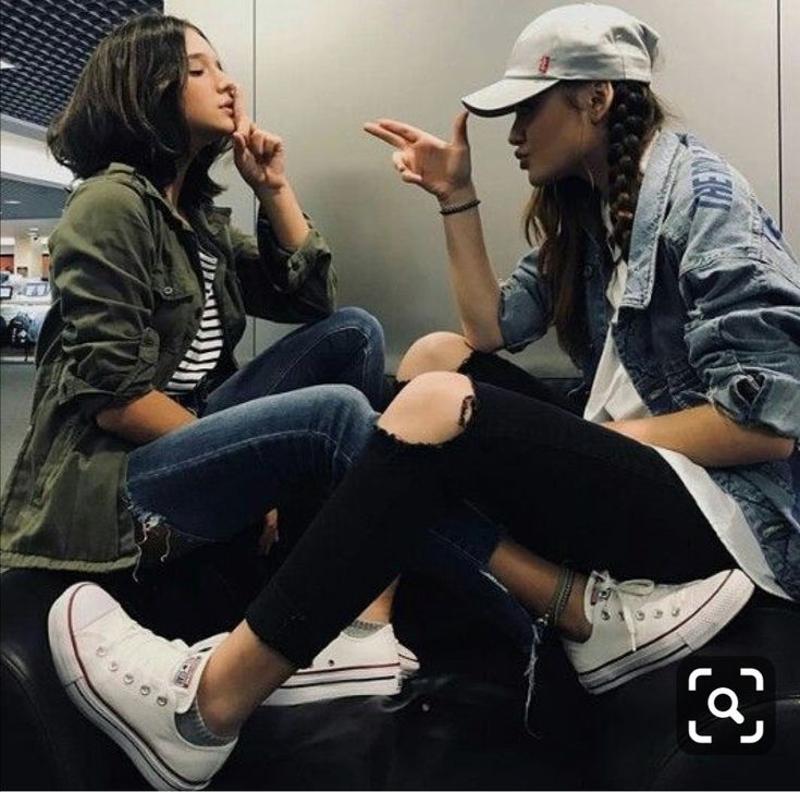 two teenage girls are hanging out together. they're both wearing trendy clothes, including denim jackets, ripped jeans, and white converse sneakers. they look happy and comfortable with each other. they seem to be best friends. the image captures the spirit of youth culture and casual style.