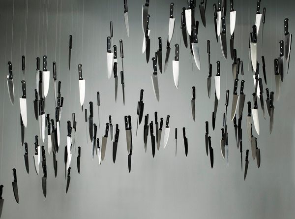 a large number of kitchen knives are hung from the ceiling against a gray wall, creating a visually striking and slightly unsettling art installation. the knives hang at various lengths, their blades gleaming in the light. the stark simplicity of the image evokes a sense of both beauty and danger.