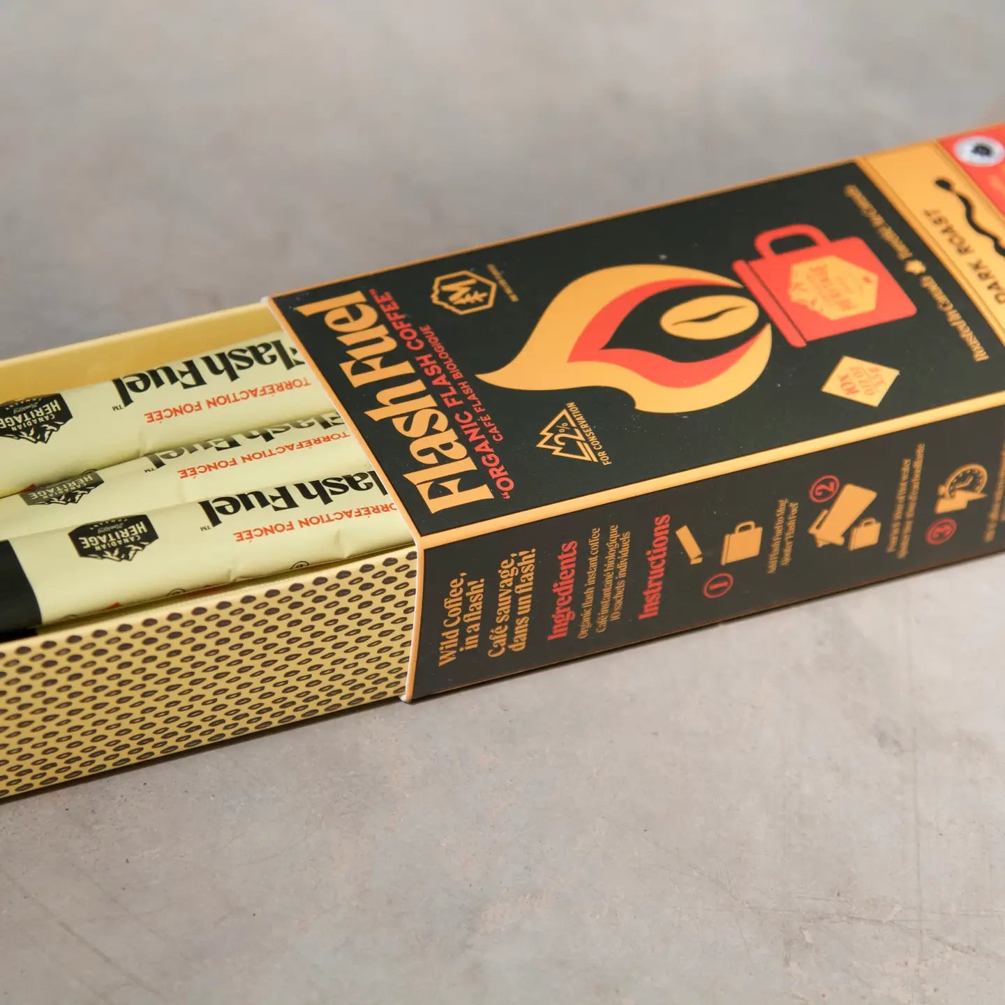 a box of flash fuel instant coffee sticks is partially open, revealing the individual packets inside. the box is black and yellow, featuring the "flash fuel" logo, a stylized flame containing a coffee bean, and the words "organic flash coffee" and "cafe flash biologique". it also includes sections for "ingredients" and "instructions" with icons. the individual coffee stick packets are yellow with the "flash fuel" logo and the phrase "torrefaction foncée". the box also indicates the coffee is a "dark roast". a "heritage" logo is visible on the coffee sticks.