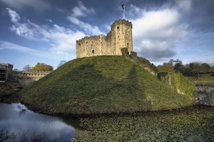 a wide shot of cardiff castle in wales, situated on a large grassy mound surrounded by a moat filled with water. the castle itself is made of gray stone and features a tall, square tower with a flag flying at the top. the sky above is blue with fluffy white clouds, and the surrounding area is lush and green with trees lining the background. the sun is shining brightly, casting long shadows across the grass and creating highlights on the water's surface. the perspective makes the castle appear grand and imposing, while the natural beauty of its surroundings adds to its charm and historical significance.