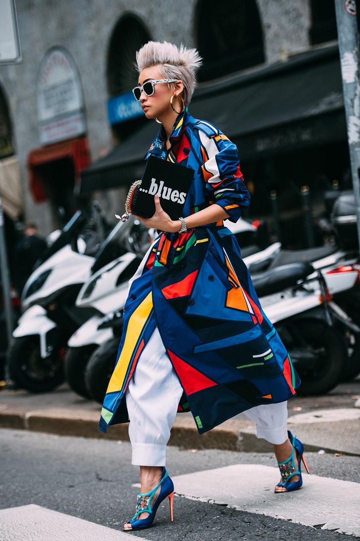 a stylish woman with short blonde hair and sunglasses walks across a crosswalk in milan, italy. she is wearing a colorful geometricprint trench coat, white pants, and blue high heels. she carries a black clutch with the word "blues" on it. the photo captures the vibrant street style of milan.