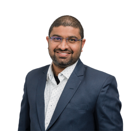 a smiling headshot of a south asian man wearing a dark navy blue suit and a white patterned shirt. he is wearing glasses and has a short, dark haircut and beard. the background is plain white. the man appears professional, friendly, and confident.  the image is welllit and highquality.
