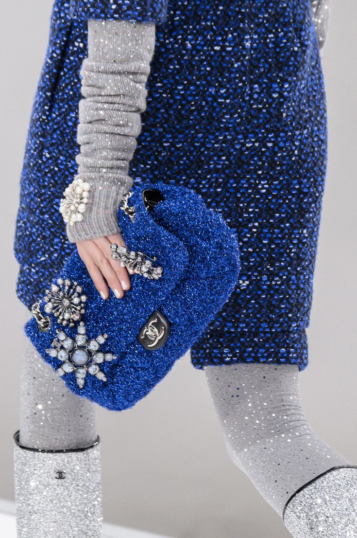 loseup view of a model's outfit showcasing a vibrant blue chanel handbag adorned with sparkling embellishments. the bag is made of a textured, possibly tweedlike material and features multiple brooches and decorative elements. the model is wearing a matching blue tweed jacket or dress, glittery silver gloves, and silver glitter boots. the image focuses on the detail of the handbag and its intricate design, highlighting the luxurious and fashionable aspects of the outfit. the background is blurred, putting all the focus on the clothing and accessories.