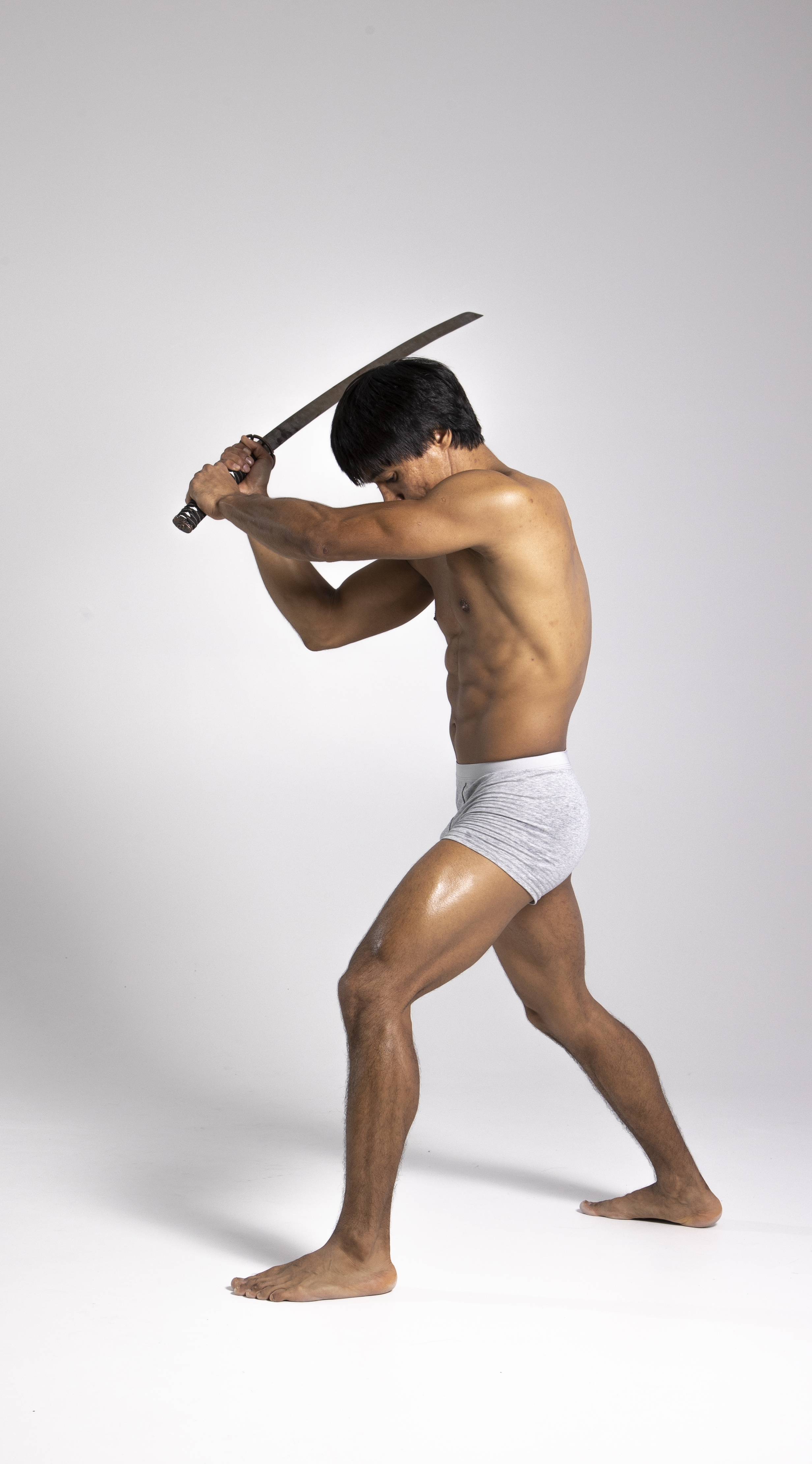 a shirtless, athletic man with dark hair and oiled skin stands in a fighting stance holding a sword. he is wearing gray underwear and is barefoot in a studio shot. his body is angled forward as he prepares to strike with the sword. the background is a plain white.