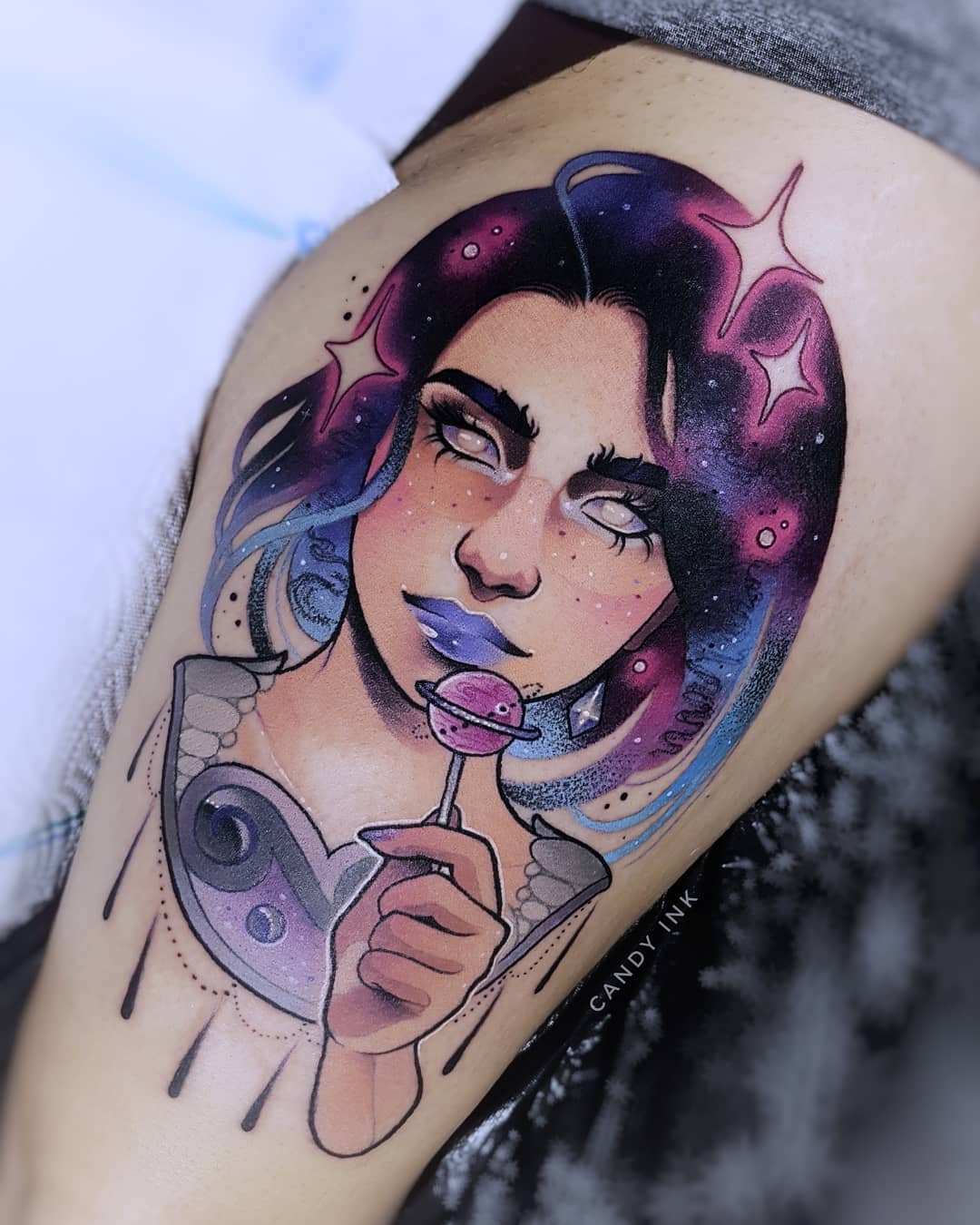 this is an image of a tattoo on someone’s upper thigh. the tattoo is of a woman with blue and purple hair that looks like a galaxy with stars in it. her eyes are white. she has a heartshaped locket around her neck, and she’s holding a lollipop that looks like a planet. the background of the tattoo is the person’s skin. the artist’s signature, “candy ink” is written in white near the bottom of the tattoo.