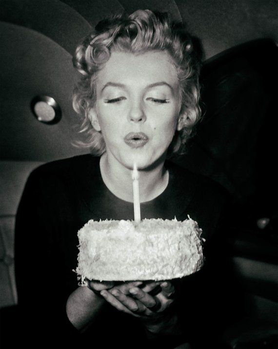 marilyn monroe is smiling as she blows out a candle on a birthday cake.  she is holding the cake in front of her with both hands.  the photo is in black and white and is likely from a photoshoot.