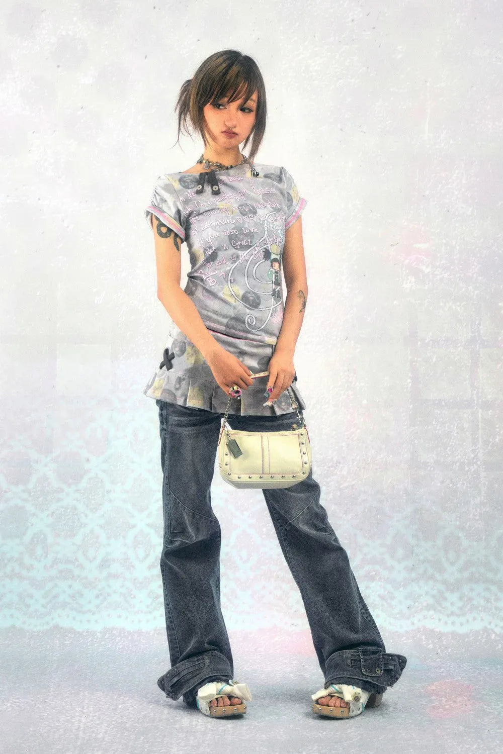 a fullbody shot of a young asian woman, possibly japanese, poses against a light, textured background. she wears a light gray graphic tshirt with a pleated hem, dark gray baggy jeans cuffed at the ankles, offwhite sandals, and a small beige handbag.  her style is casual and incorporates elements of early 2000s fashion. she is wearing a simple necklace. her expression is somewhat downcast, and her pose is relaxed but somewhat formal. the image has a soft, slightly muted color palette.