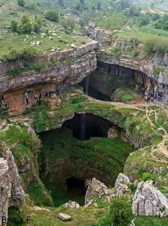 the image depicts a stunning natural sinkhole known as the baatara gorge waterfall in lebanon. the landscape features three natural bridges stacked on top of each other within the sinkhole's depths. water cascades down, forming a waterfall that plunges into the cavelike base. the sides of the sinkhole are characterized by layered rocks and lush green vegetation clinging to the cliffs. the surrounding mountainous terrain is visible in the background, creating a scenic outdoor view. people standing near the edge give an idea of the scale and grandeur of the natural formation.