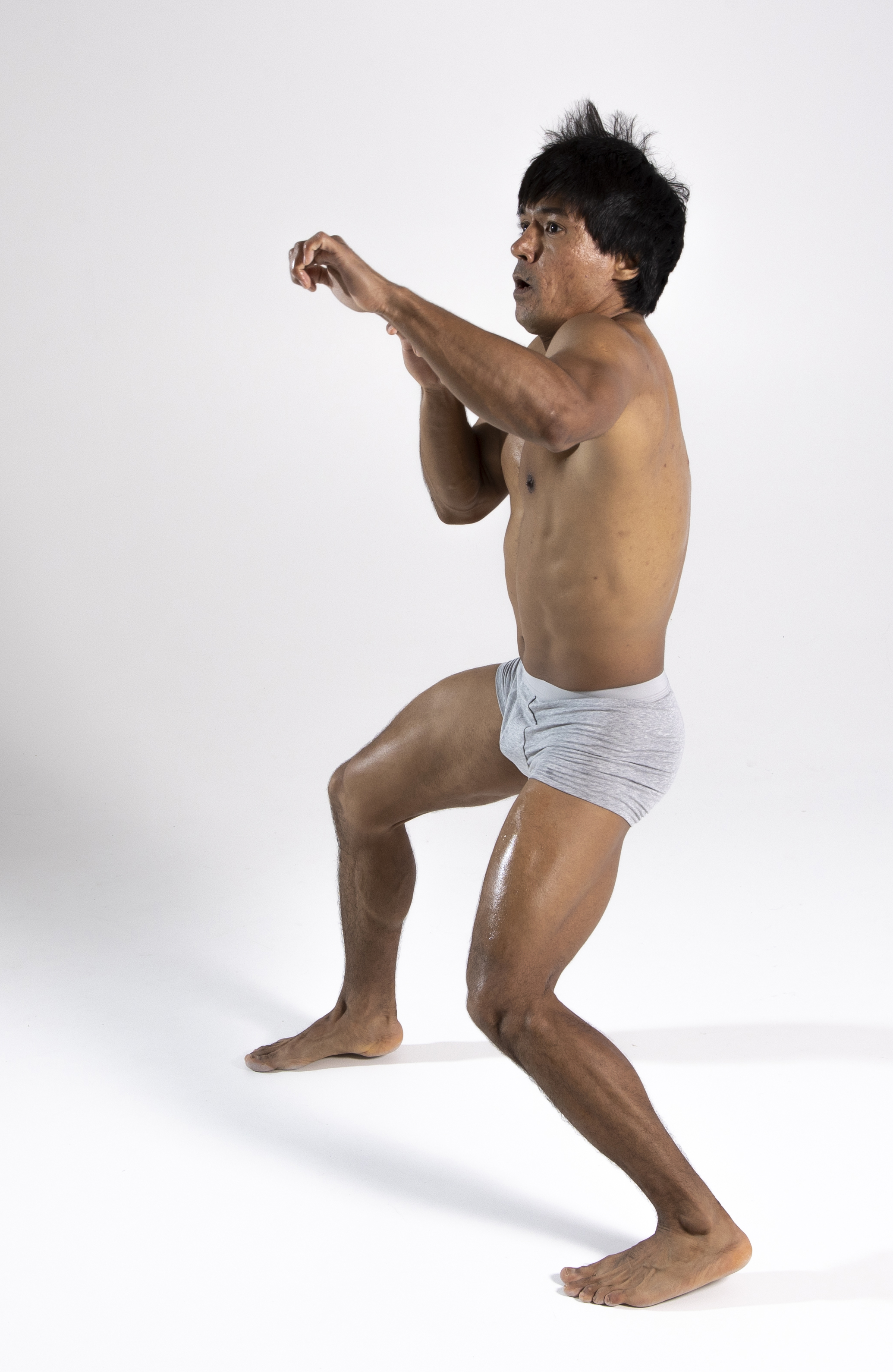 a shirtless, muscular man wearing underwear is posing in a boxing stance against a white background. he has a defensive stance, with his fists raised and a look of concentration. he is barefoot and appears sweaty, suggesting intense activity.