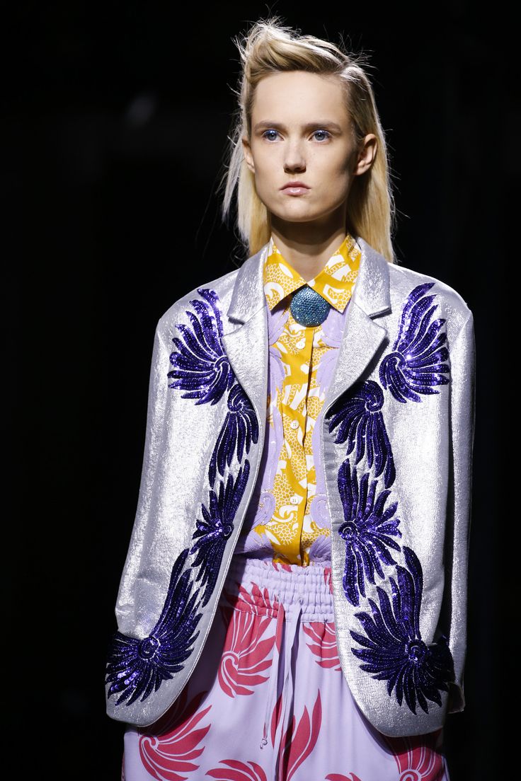 a fashion model with blonde hair styled in a high quiff, is showcasing a silver sequined blazer adorned with purple floral embroidery.  underneath, she wears a lavender and yellow patterned shirt and matching lavender pants with a pink floral design.  she is also wearing a turquoise brooch. the model's expression is serious and she is posed against a black background. the overall style appears to be high fashion and contemporary.