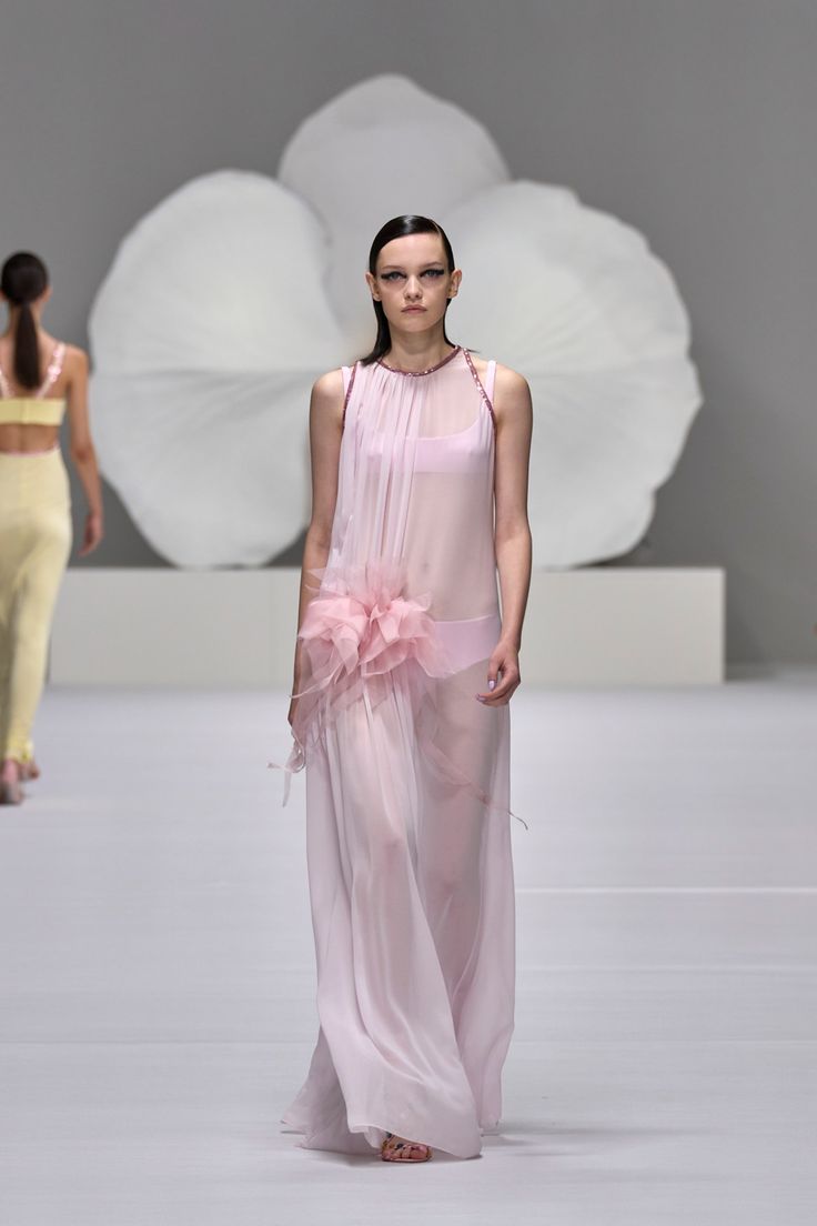 a model walks the runway during a fashion show, wearing a long, sheer pink dress with a large pink flower detail on the side. she is wearing a matching pink undergarment. the backdrop is a clean white space with a large, cloudlike structure behind her. another model, wearing a yellow ensemble, is walking slightly ahead of her, partially out of focus.