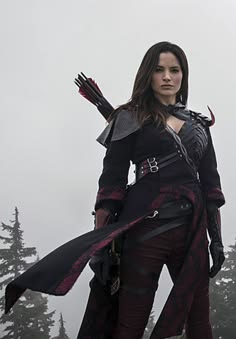 a woman is wearing a black and red hunting outfit. she is holding a bow and arrow and is looking seriously towards the camera.  the fog and trees surrounding her suggest she is in a forest.