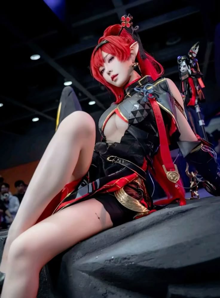a woman cosplaying a character with red hair and elf ears sits on a dark surface, likely a prop. she is wearing a black and red costume with a lowcut neckline. there is a figurine of a character behind her. the scene is likely at a convention.