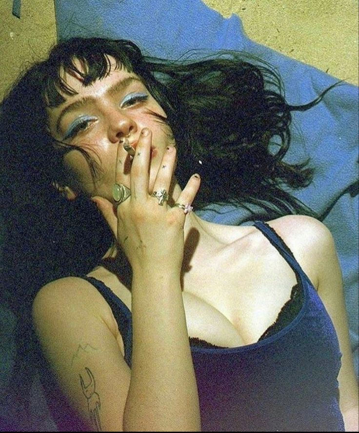 a close up portrait of a young woman with dark hair and blue eyes. she is wearing a blue tank top and has a cigarette in her mouth. she is lying down and looking at the camera. the image is in focus and the colors are vivid. the woman's expression is relaxed and she appears to be enjoying herself. the background is blurred out of focus.
