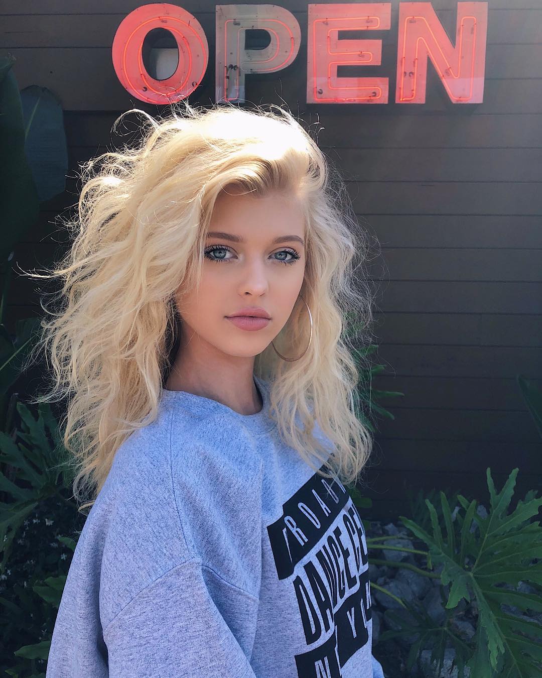 a young woman with blonde wavy hair and blue eyes is standing outside in front of a wall with a red neon "open" sign. she is wearing a gray sweatshirt with a black graphic on it, and large hoop earrings. green leafy plants are visible in the background. the lighting suggests it is daytime.
