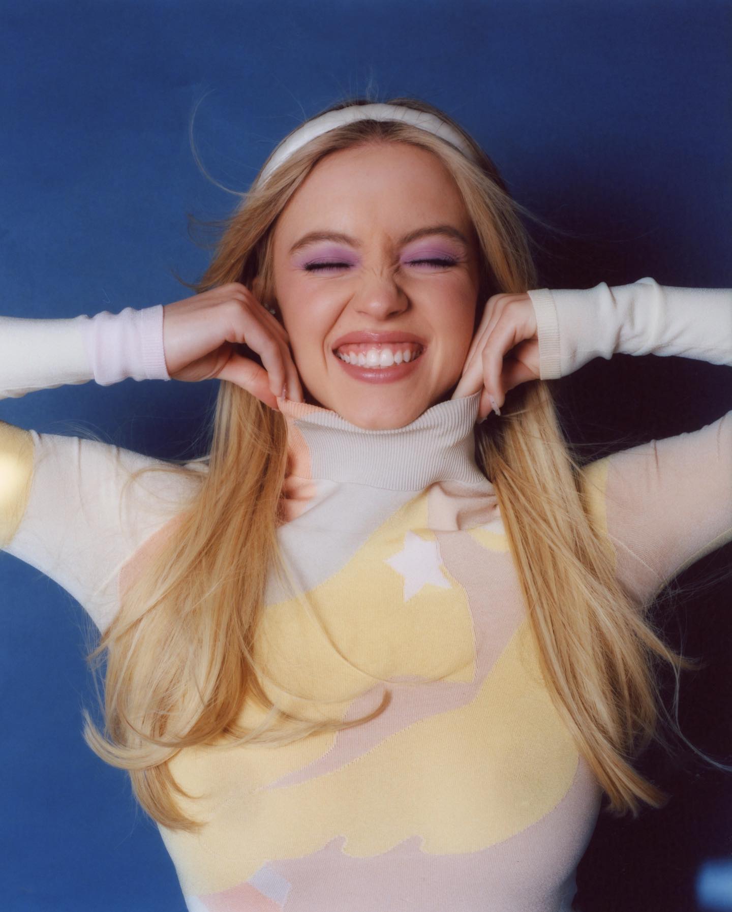closeup portrait of a young blonde woman with long hair smiling brightly. she is wearing a pastel yellow and pink turtleneck sweater with a subtle star design, a white headband, and subtle purple eyeshadow. her expression is joyful and carefree. the background is a solid blue. the image has a soft, slightly dreamy quality.