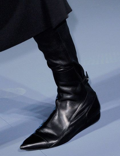 a closeup of a model's legs wearing black, pointytoe, flat boots. the model is wearing a black outfit, and the boots are made of leather. the boots are a very sleek and stylish design.