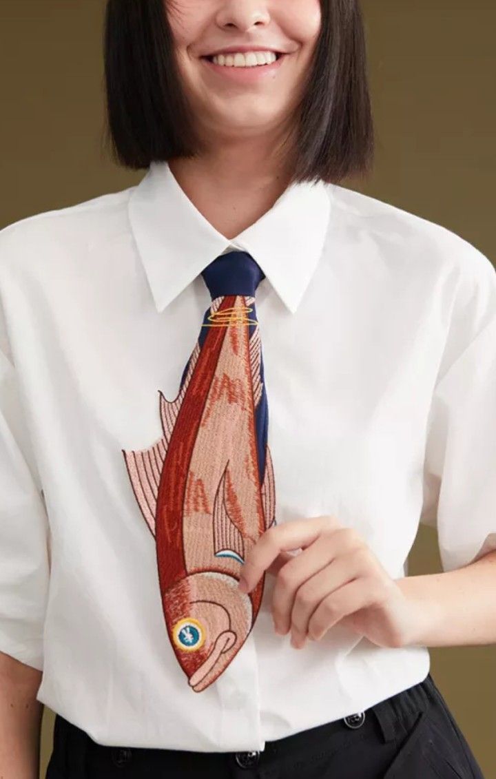 a closeup portrait of a woman with short dark hair wearing a white buttondown shirt with a fish embroidered on the tie. she is smiling at the camera.