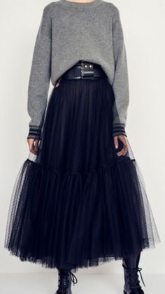 a woman is posing in a studio wearing a grey sweater and a black tulle skirt. she has a black belt around her waist, and black boots on her feet.