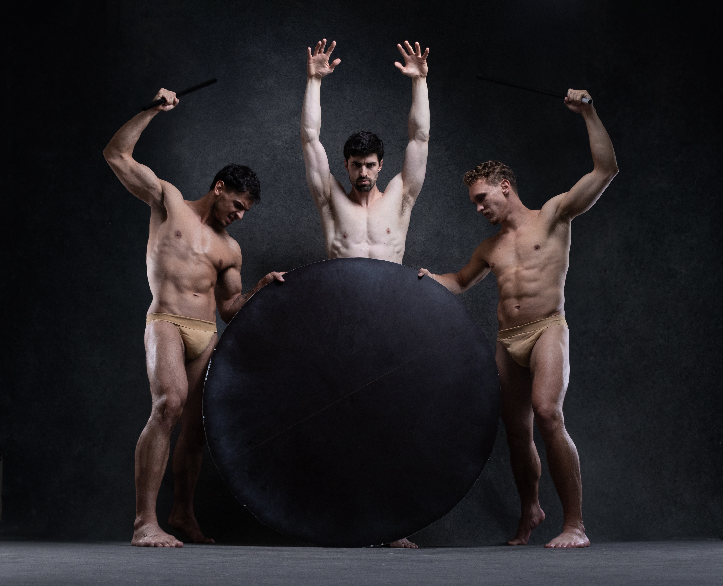 three muscular men wearing only beige thongs pose with their bodies in a dance stance. the man in the middle stands behind a large black circular object and has his arms above his head, palms facing forward. the men on each side of him are facing towards him. each man has one arm raised above his head holding a black stick and the other arm bent, holding onto the large black circular object.