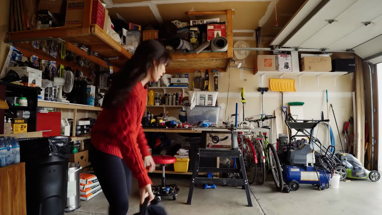 the image shows the inside of a cluttered garage.  a young woman in a red sweater stands in the center, seemingly about to pick up a black bag from a rolling stool. the garage is filled with various tools, equipment, and storage solutions, including wooden shelves, metal racks, and plastic bins. the space appears somewhat disorganized, with items scattered across the floor and workbench. the garage door is open, letting in natural light that illuminates the scene.