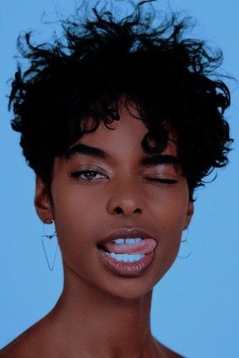 a young black woman with curly black hair and a playful smile. she is wearing a pair of silver triangle earrings and has on dark eyeliner.  her skin tone is warm and she is looking directly at the camera. the backdrop is a solid blue, with the woman taking center stage.