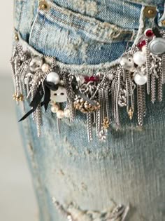 a closeup shot of a pair of distressed denim jeans embellished with silver chains, beads, and other metallic accents. the jeans have a vintage, grunge aesthetic, and the embellishments add a bold, edgy touch. the fabric is frayed and ripped, creating a unique texture. the image captures the details of the jeans, highlighting the intricate work that has gone into their design.
