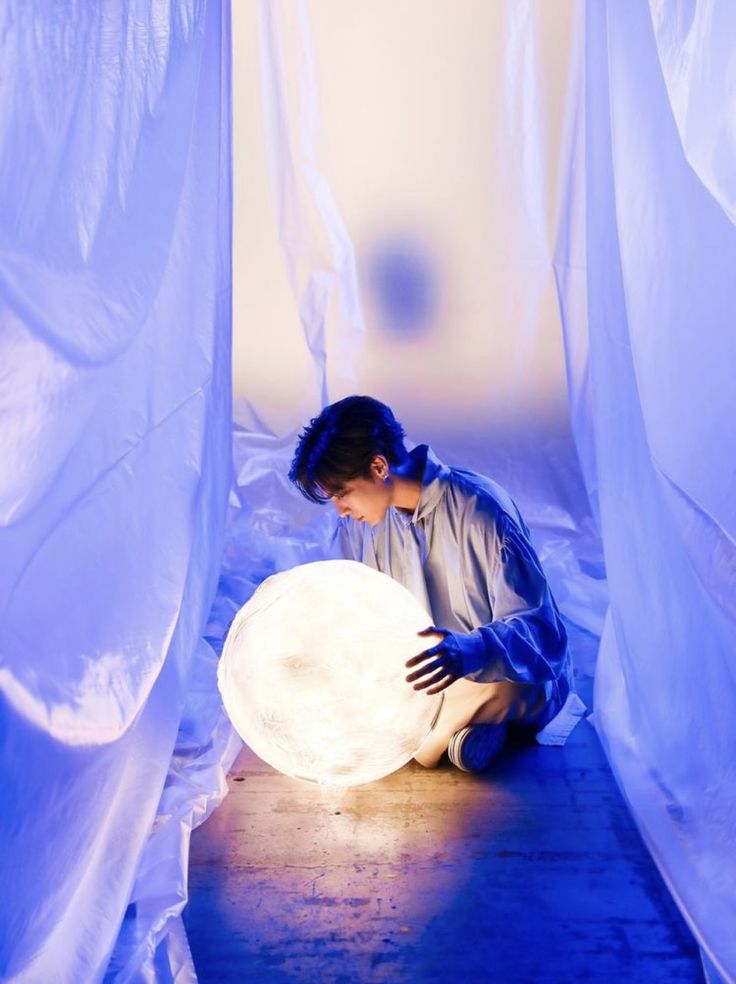 a young man sits on the floor in a dimly lit studio space, bathed in the soft glow of a large, round light source he holds.  blue plastic sheeting drapes the background and sides, creating a sense of depth and otherworldliness. the overall mood is serene and contemplative.