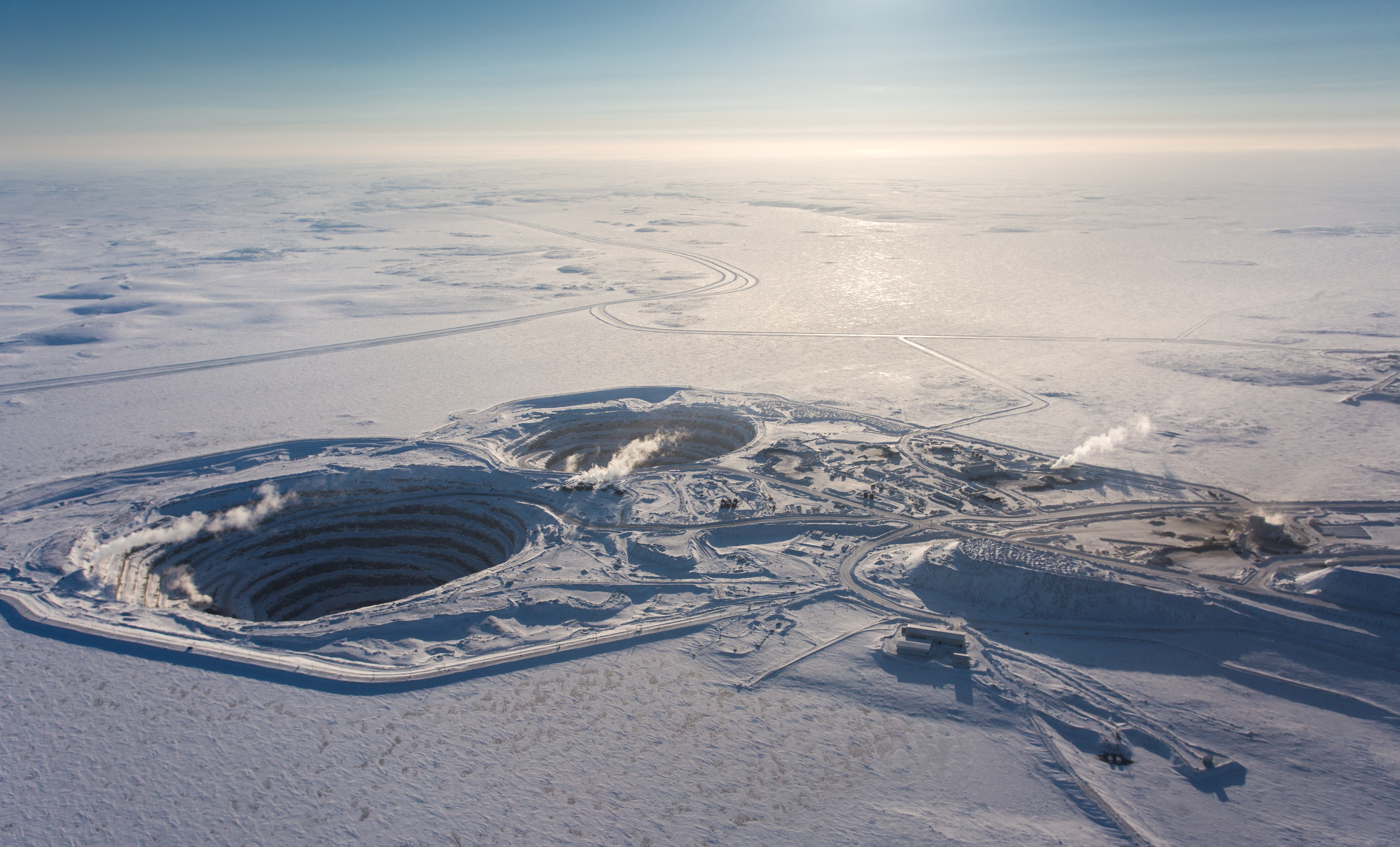 a bird's eye view of a massive diamond mine in the snowy arctic landscape. the vast open pit mine is visible with its multiple layers and a large crater in the middle. the mine appears to be in a remote and isolated location, surrounded by white snow and ice. there is a network of roads and infrastructure surrounding the mine, suggesting a largescale mining operation. this is a captivating visual of the impact of human industry on the natural environment.