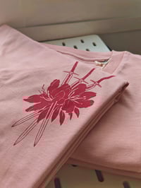 a closeup shot of a folded pink tshirt featuring a graphic design of three swords intersecting within a stylized red flower. the tshirt is soft and has a casual style. the image showcases the design and texture of the fabric. the tshirt is neatly folded and layered, suggesting a display or merchandise context.  the background is blurred, drawing attention to the tshirt itself.