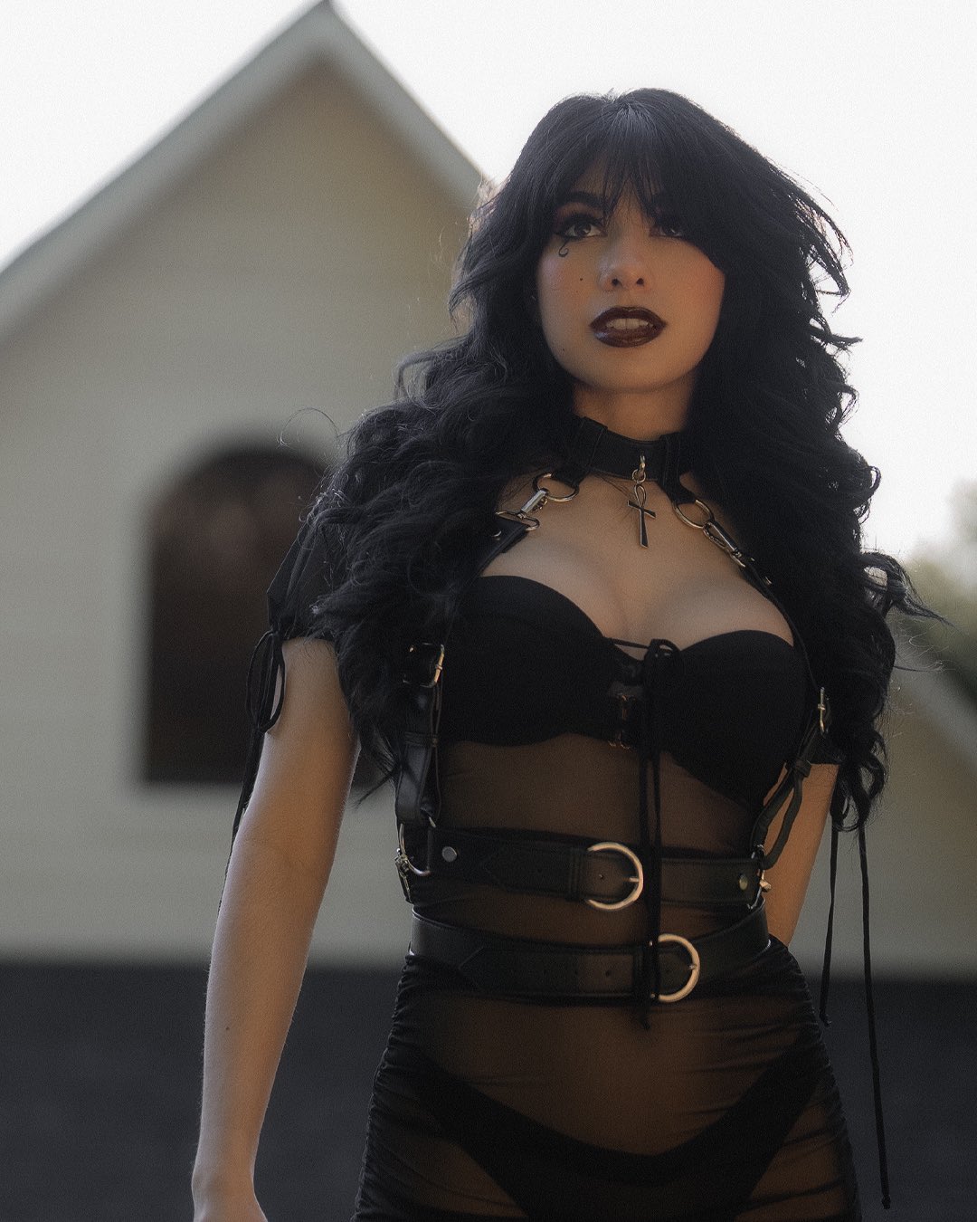a woman dressed in goth style. she is wearing a black dress with a black harness. she also has a black necklace with a cross. she is wearing dark makeup and her hair is styled in a dark, wavy manner. she is standing outdoors with a building in the background.