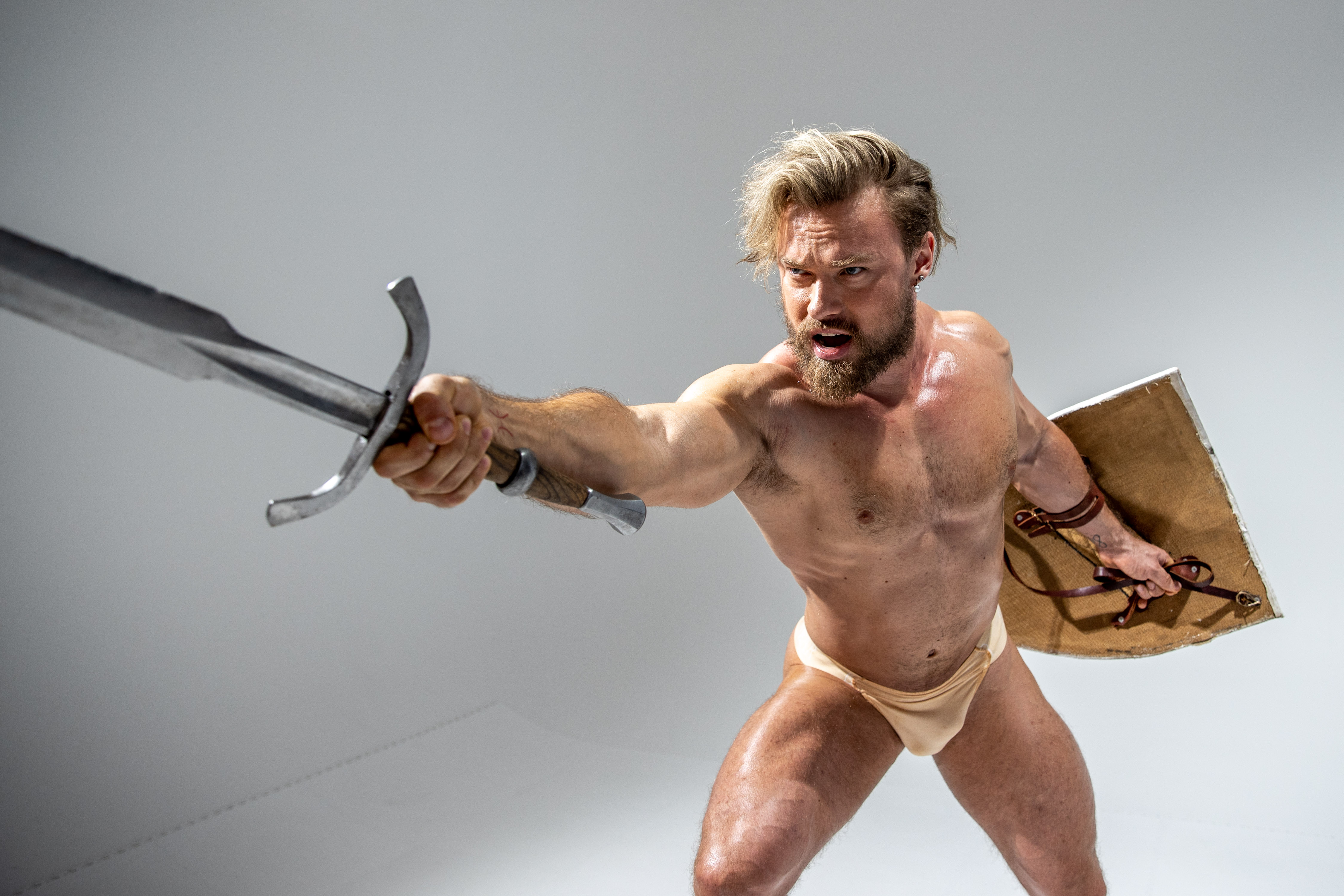 a muscular man with a beard and blonde hair stands in an attack position, brandishing a sword and shield. he wears only beige underwear and a leather wrist cuff. the photo is taken from a low angle, with bright studio lighting.