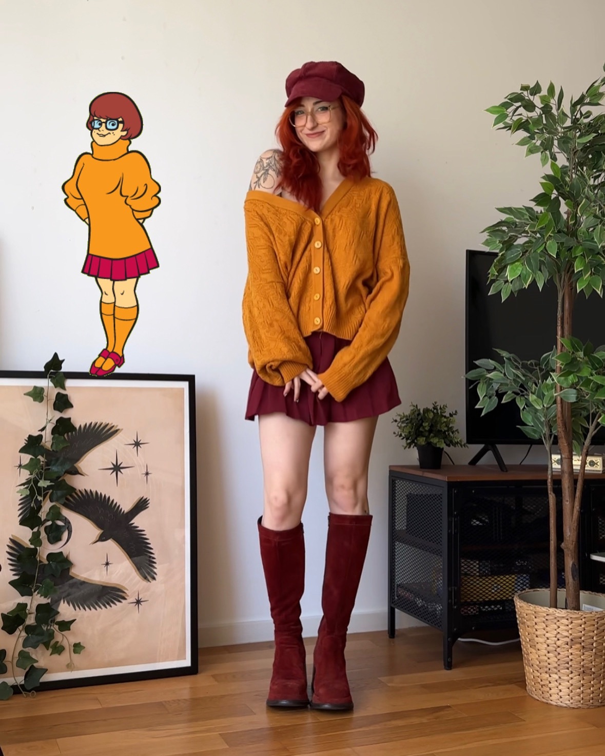 a fulllength photograph shows a young woman cosplaying as velma dinkley from scooby doo. she's wearing a mustard yellow cardigan, a maroon aline skirt, and maroon kneehigh boots.  she has red hair styled in a similar manner to velma's, and is wearing glasses and a newsboy cap.  a stylized illustration of velma is visible on the wall behind her. the setting appears to be a living room with plants, a framed print on the wall, and dark furniture.  the overall style evokes a retro or 70s vibe.