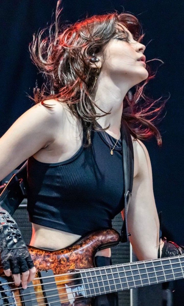 a woman with brown hair is playing a bass guitar on stage. she's wearing a black tank top, a necklace, and earrings. she has gloves on her hands and is focused on playing the instrument. the stage lights illuminate her and the guitar.