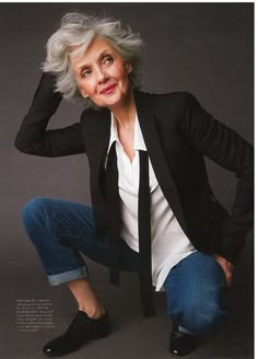 a woman in a black blazer, white shirt, and denim jeans poses in a studio. her gray hair is styled in a tousled look, and she is wearing black shoes.