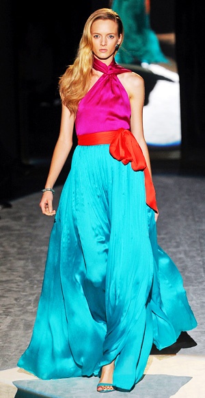 a model walks down a runway wearing a pink halter top, a turquoise pleated skirt, and a red sash.