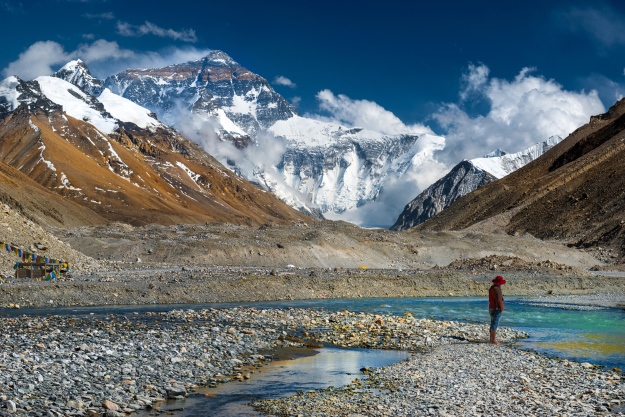 the image shows a landscape with a large snowcovered mountain in the background, possibly mount everest. in the foreground, there is a wide, rocky riverbed with turquoise water flowing through it. a person wearing a red hat stands on a small rocky bank, looking out at the scenery. the sky is blue with scattered white clouds. the valley is surrounded by steep, rocky mountains, some with patches of snow.