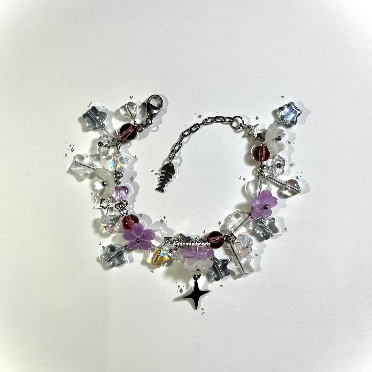 a closeup shot of a beaded bracelet featuring a variety of charms and beads. the bracelet includes clear beads, purple flower beads, starshaped beads in various colors, and a small fish charm. it is held together by a silver chain and fastens with a lobster clasp. the beads and charms are arranged seemingly randomly along the bracelet.