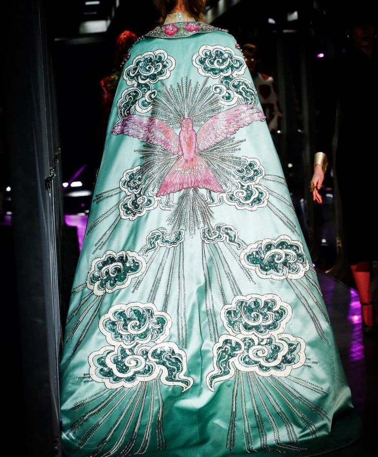 back view of a light blue gown or cape adorned with a pink dove and cloud embellishments, accented by sequins and embroidery, likely on a runway.