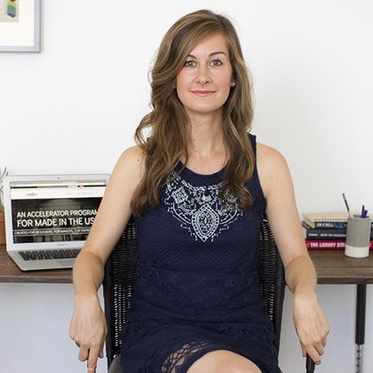a woman is sitting in a chair at a desk. she is wearing a sleeveless, lace dress. there is a laptop with an "accelerator program" message on the screen, some books, and a pencil holder on the desk.