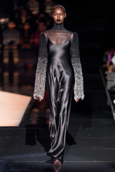 a black woman, walking the runway wearing a long black silk dress,  with long silver sleeves and a silver choker. the dress is a long black silk gown, with a loose fit and a vneckline. the model has black lipstick and black hair pulled back away from her face. she is walking on a black runway, in front of a crowd of spectators. the model is giving an intense look to the camera.