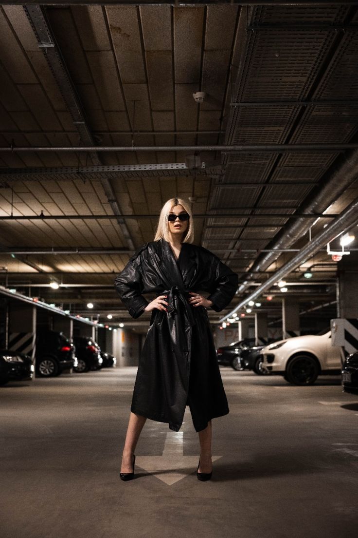 a blonde woman in a black leather trench coat and sunglasses poses confidently in a dimly lit parking garage.  she stands with her hands on her hips, her posture exuding selfassuredness. cars are parked in the background, adding to the urban setting. the overall mood is dark and stylish.
