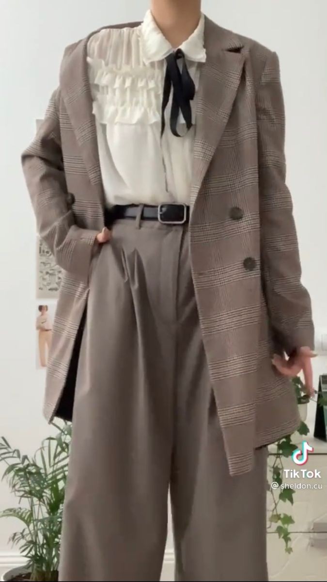 a person is wearing a stylish outfit consisting of a plaid blazer, a white blouse with ruffles and a black ribbon tie, wideleg gray pants, and a black belt. the person's hand is in the pocket of their pants. the overall look is chic and puttogether, indicative of a fashionforward style. there are plants in the background.