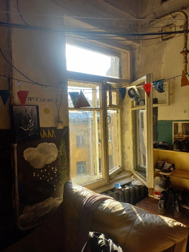 a photograph of a room in an old building, bathed in sunlight streaming through a large, aged window. the room is filled with various objects, including a worn couch, shelves with shoes, and walls adorned with graffiti, drawings, and string lights with small flags. the overall atmosphere is vintage and somewhat cluttered, with a mix of textures and colors creating a bohemian feel. the sunlight highlights the textures of the walls and the wornout furniture. the scene suggests a livedin space, reflecting the character and history of the dwelling.
