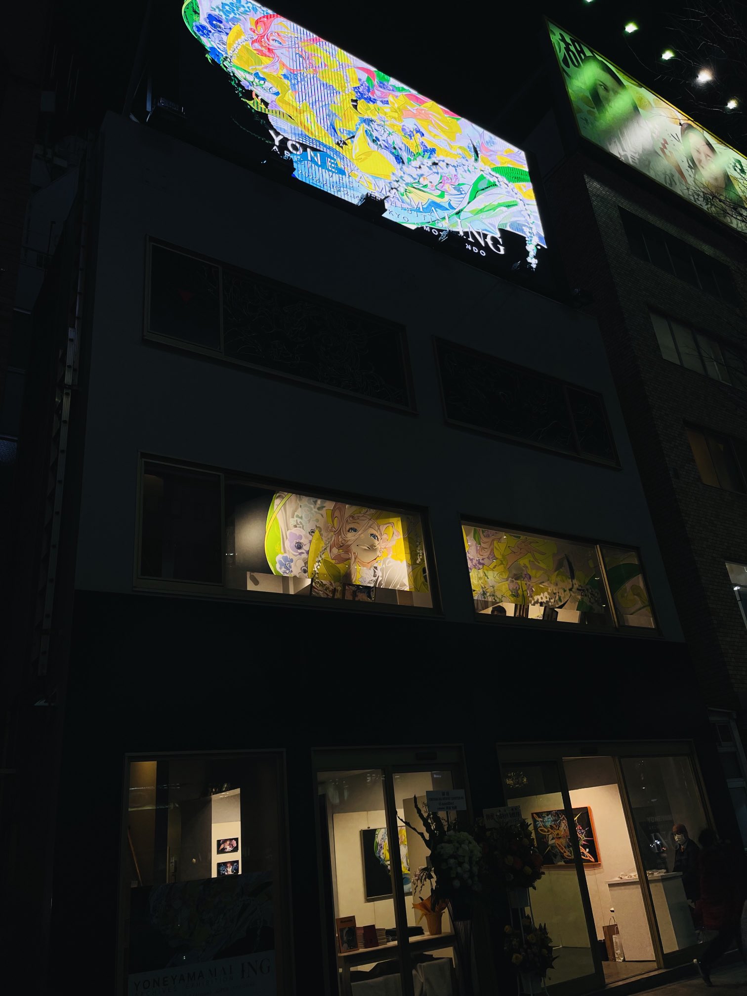 a photograph of a brightly lit art gallery at night. the building has multiple levels showcasing different artworks, including a large billboard on the top displaying colorful abstract art. the windows on the second level feature portraits while the ground floor exhibits colorful paintings. the overall ambiance is vibrant and lively, indicative of a bustling city nightlife.