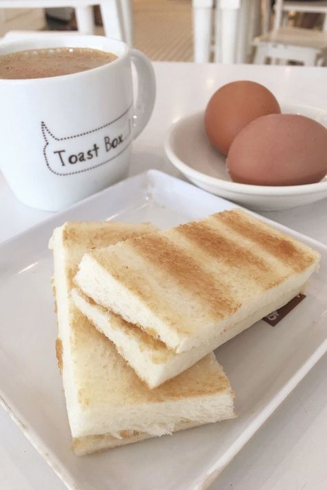 a closeup shot of a plate of kaya toast and softboiled eggs, accompanied by a mug of coffee. the mug has the "toast box" logo on it. the setting appears to be a cafe or restaurant. the food is presented simply but elegantly on white plates and a white table. the overall tone of the image is bright and airy.