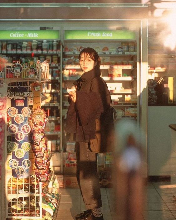 a young woman stands in the brightly lit aisle of a convenience store, bathed in the warm glow of the setting sun.  she's dressed in casual streetwear, browsing the shelves stocked with colorful snacks and drinks.  the image has a candid, filmlike quality with a focus on capturing a fleeting moment of everyday life.