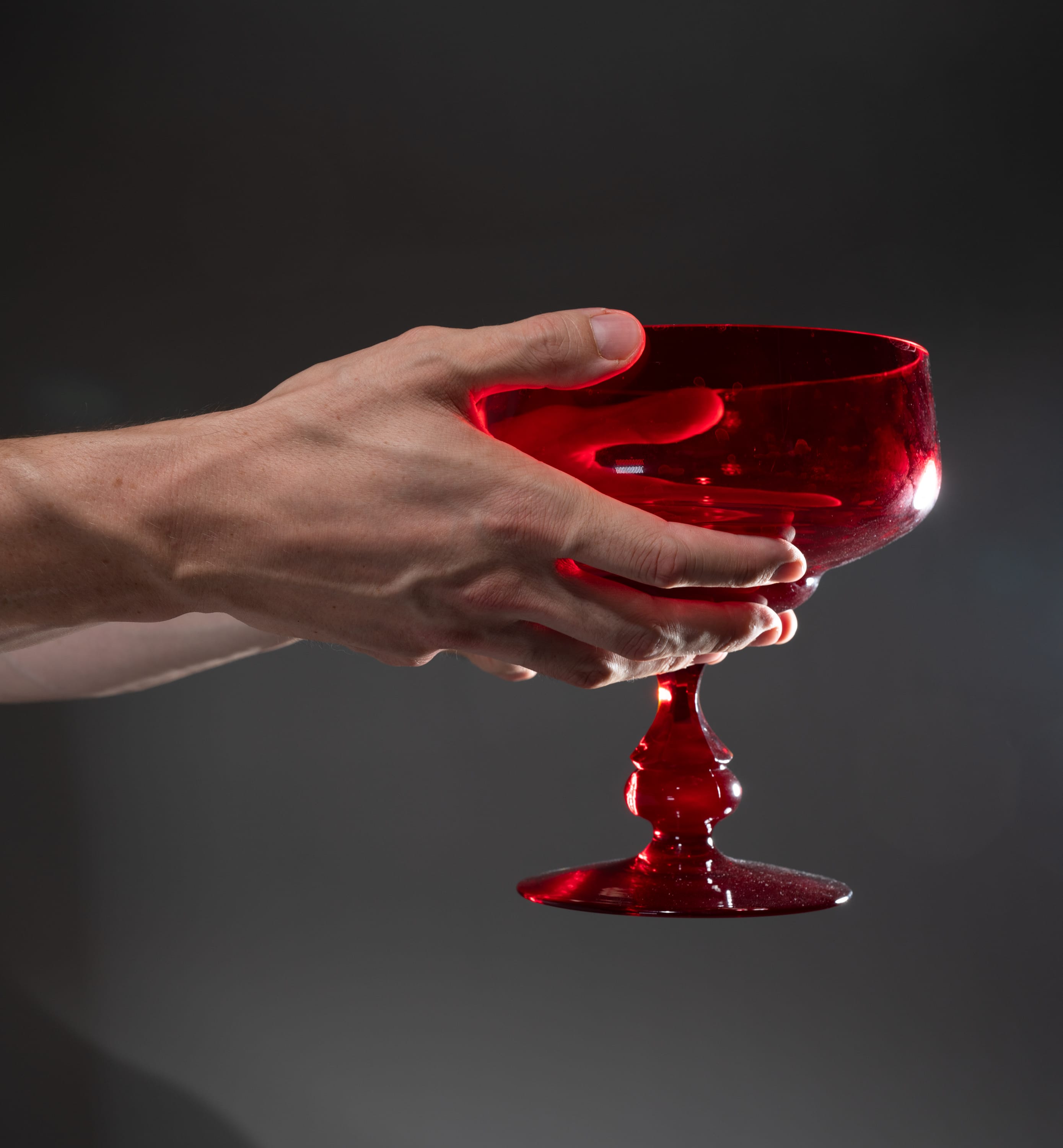 a closeup studio shot of a man's hand holding a red glass goblet against a dark gray background. the goblet is empty. the man's hand is positioned in the middle of the glass, gripping it firmly. the lighting is dramatic, highlighting the curves of the glass and the contours of the hand.